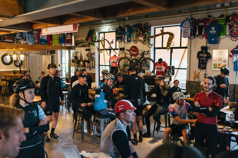 Helmeted riders drink coffee under hanging jerseys inside the Cyklist café in Eindhoven before the Backyard Breakaway.