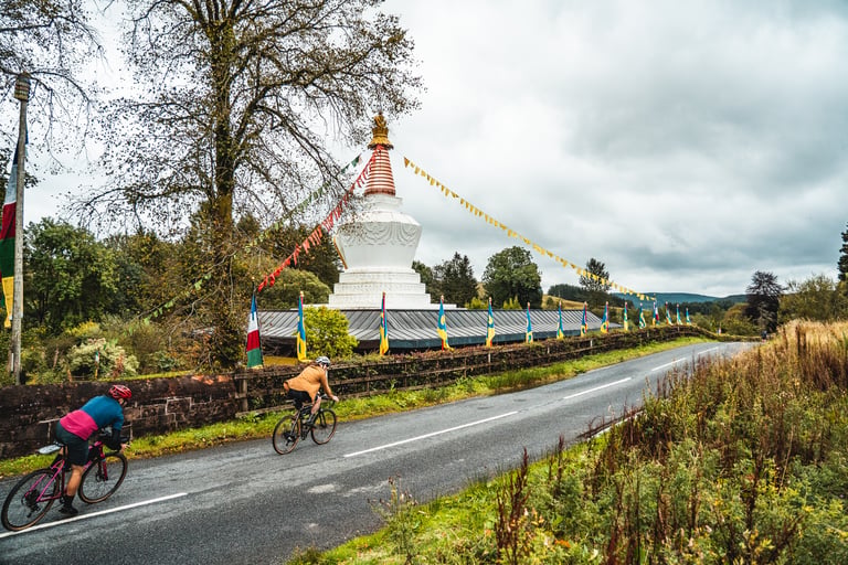 Two riders pass a white Buddhist stupa hung with prayer flags on a wet road during the Frontier 300 recce.