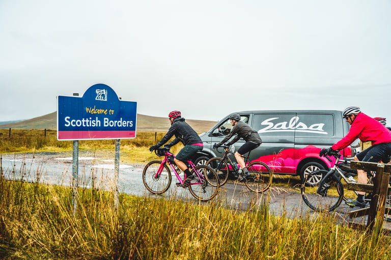 Riders on gravel bikes pass a Salsa van parked beside a Welcome to Scottish Borders road sign in the rain.