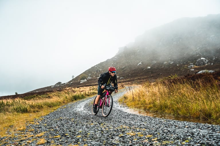 Tom of Lyon Outdoor rides the hot pink Salsa Journeyman along a wet gravel track below a misty crag.