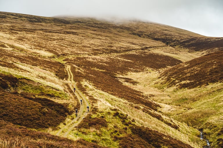 Four riders follow a twin track through a vast moorland valley under low cloud on the Frontier 300 route.