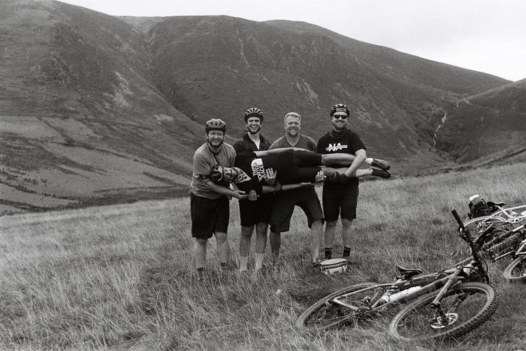 Black and white film shot of four riders holding a fifth horizontally in the Lake District hills, bikes on the grass.