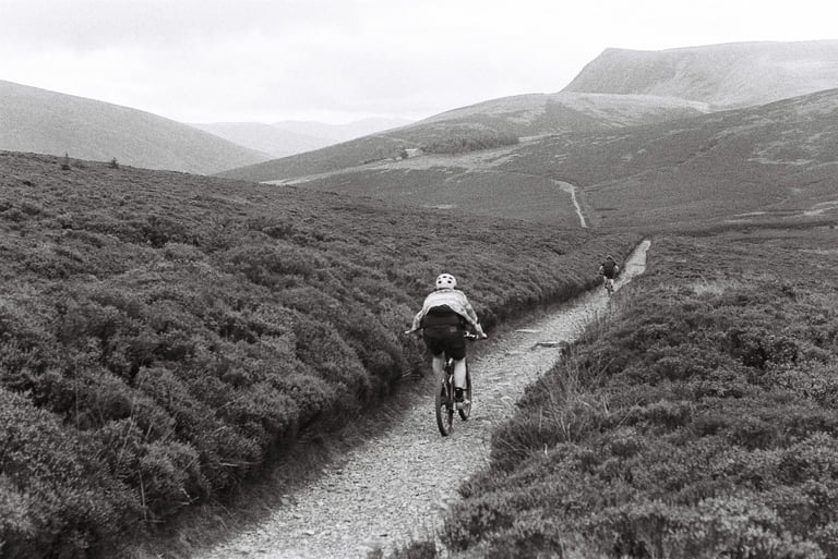 Two riders follow a narrow stony track through heather towards the Lake District fells, in black and white.