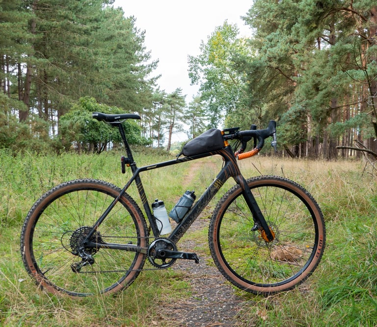Salsa Warroad on knobbly 650b tyres with a top tube bag, parked on a grassy track through pine woods.
