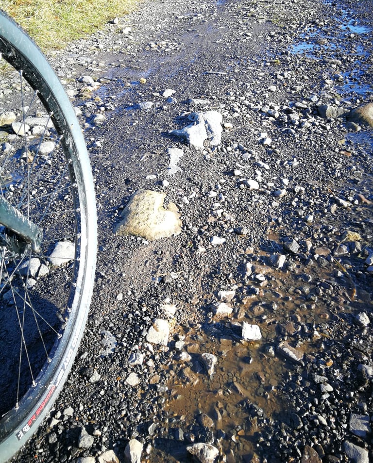 Front wheel fitted with a Vittoria Terreno Dry tyre beside a wet, stony gravel track with puddles.