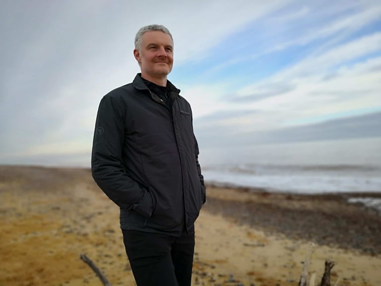 Rider wearing the Endura Hummvee Shacket dark windproof side out, hands in pockets on a windswept beach.