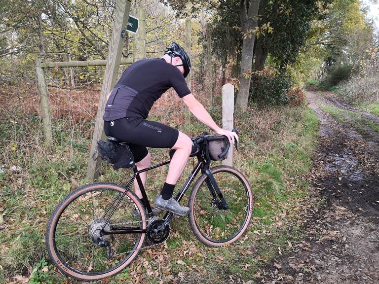 Rider in black Endura MTR Spray bib-shorts and jersey pauses on a loaded gravel bike beside a muddy woodland bridleway.