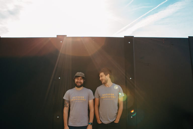 Outdoor Provisions founders Christian and Luke laughing together in matching branded T-shirts against a dark wall.