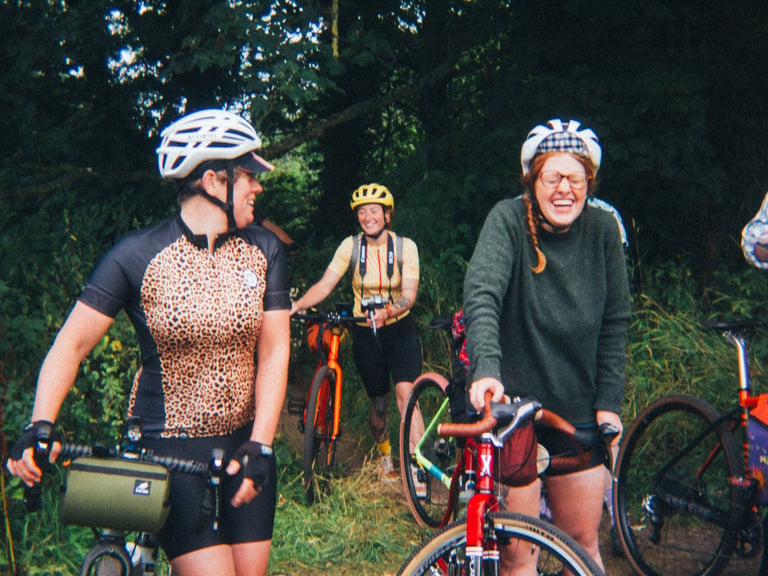 Laughing riders with gravel bikes and bar bags gather on a woodland track at the Low Key Lakes Weekender.