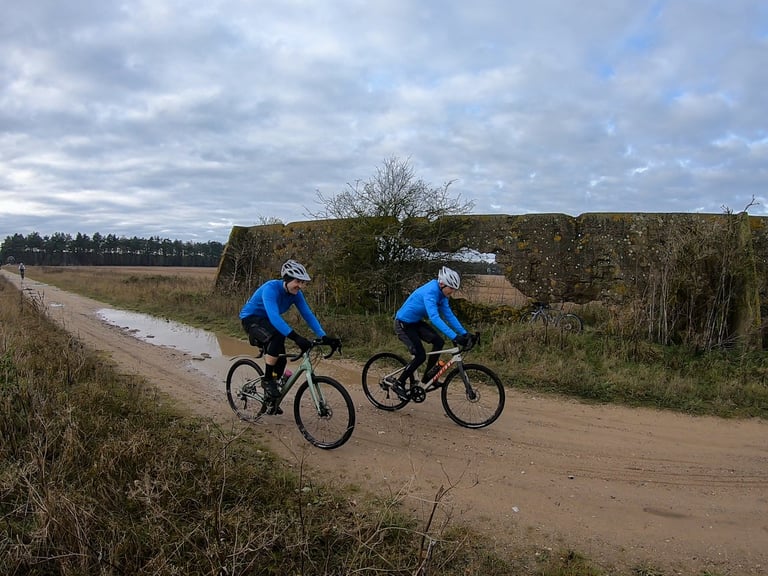 Two riders in blue jerseys on gravel bikes pass a lichen-covered concrete wall on a sandy track in Kings Forest, Suffolk.