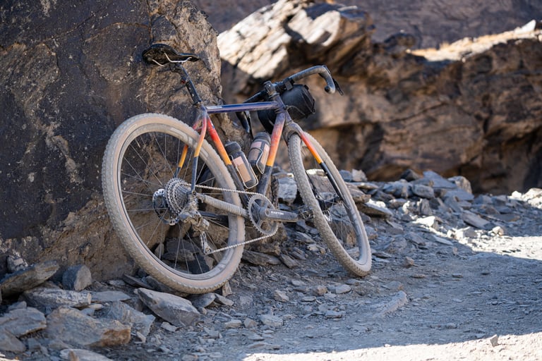 Dusty gravel bike with a bar bag and bottles leans against a rock face beside a rocky track in Tajikistan.
