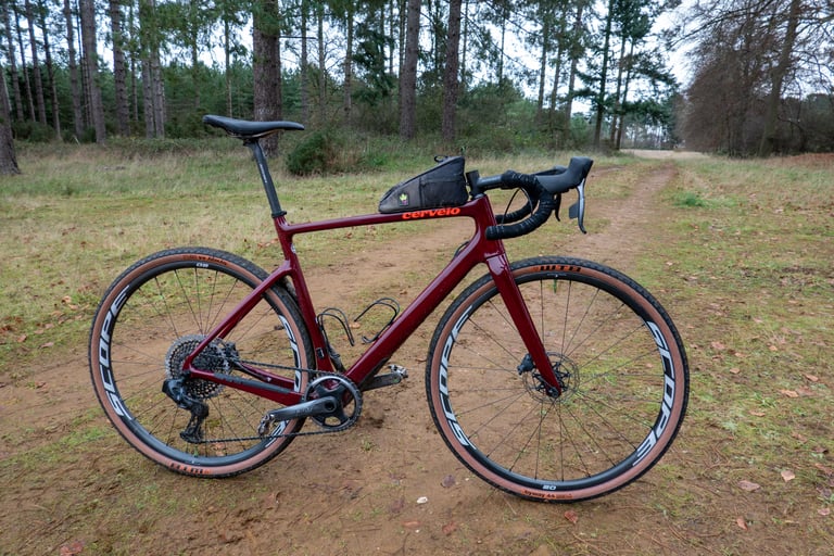 Burgundy Cervelo Aspero with SRAM AXS and a top tube bag parked on a sandy track among pine trees.