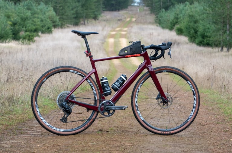 Cervelo Aspero gravel racer with top tube bag and two bottles, side view on a grassy forest track.