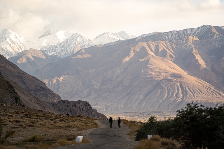 Two riders on a dirt road below snow-capped peaks in the Pamir Mountains, Tajikistan, in evening light.