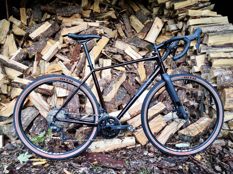 Side view of the Pinnacle Arkose D3 gravel bike with WTB tyres, propped against a stack of chopped logs.