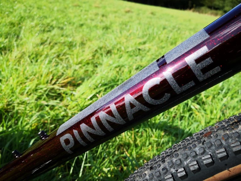 Close-up of the Pinnacle logo on the Arkose D3 frame, red glitter flecks catching the sun in the deep red paint.