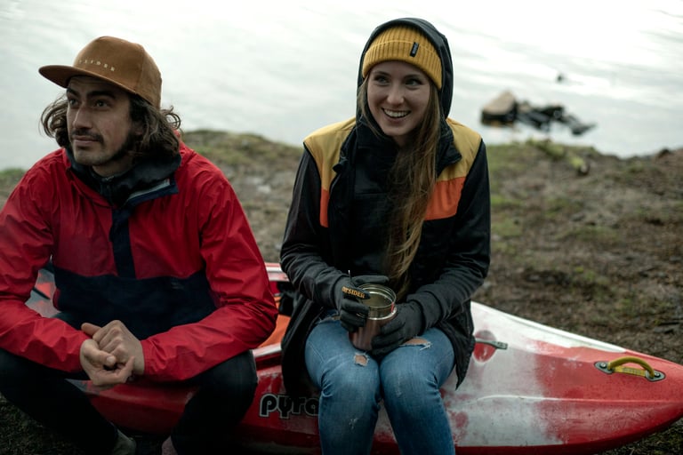 Two people in The Overland outdoor clothing sit on a red kayak at the water's edge, one holding a steel mug.