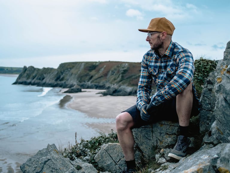 Rider in a checked Overland shirt and cap sits on coastal rocks, looking over a bay and sandy beach.