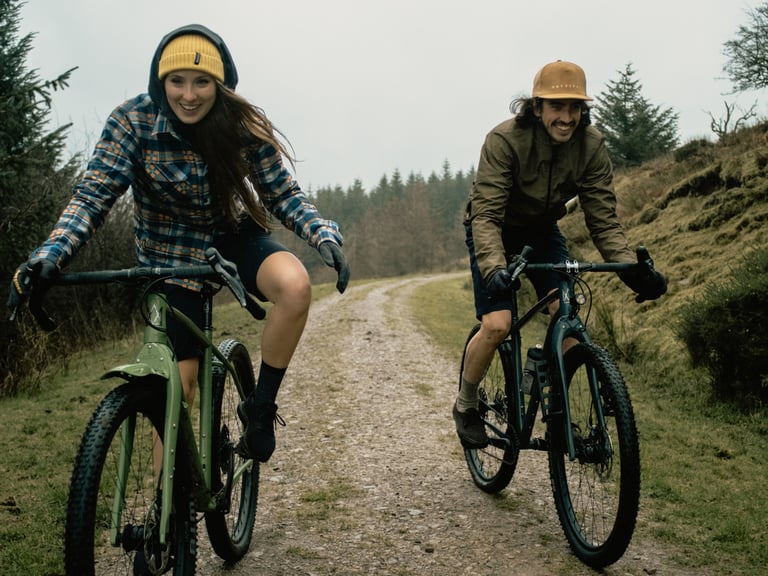 Two riders in The Overland clothing smile as they ride bikes along a gravel track through forest.