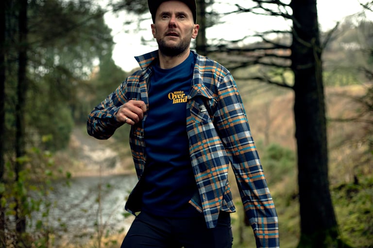 Walker in a Morvélo Overland T-shirt and checked overshirt strides through lakeside woodland without a bike.