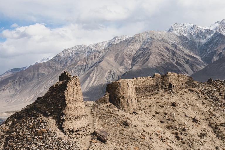 Ruined stone fortress towers on a ridge with snow-dusted mountains behind, on The Service Course's Tajikistan route.
