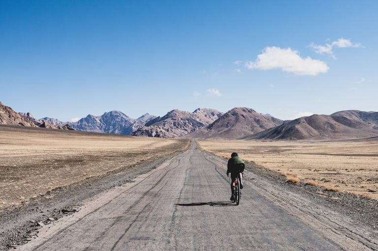 Lone rider on a cracked, empty highway crossing a high plateau toward mountains in Tajikistan's Pamirs.