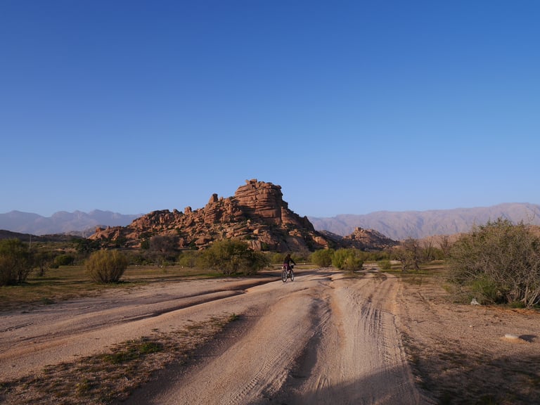 A rider on a sandy track heads towards a rocky outcrop with mountains behind, in Morocco's Atlas Mountain Race country.
