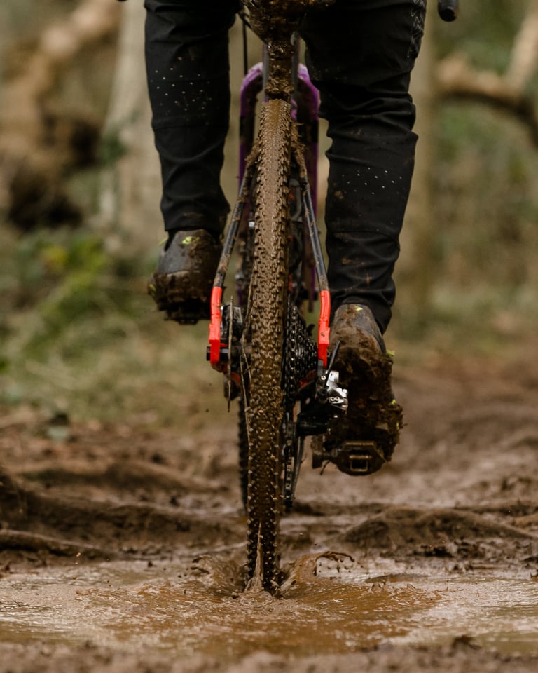 Rear view of a rider on a WTB Raddler tyre, splashing through a muddy puddle on a wooded trail.