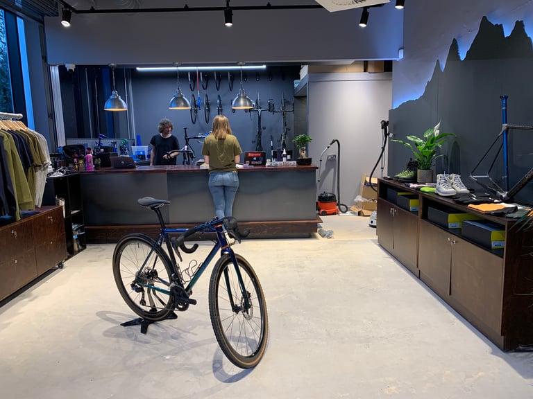 Shop floor of The Service Course Wilmslow with a bike on a stand, the workshop counter, clothing rails and a frame.