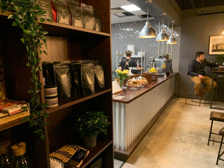 La Fabrica coffee bags and chamois balm on a shelf, with the cafe counter and a customer beyond, in Wilmslow.