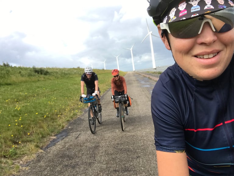 Selfie of a rider in a patterned cap with two friends on bikepacking bikes behind, on a path past wind turbines.