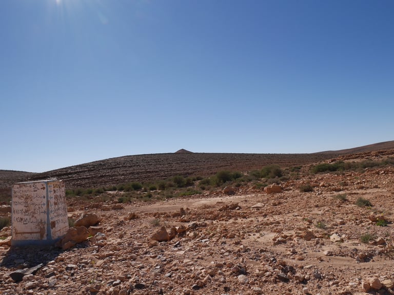 A hand-painted roadside marker block lists towns on a barren, stony Moroccan plateau under a clear sky.