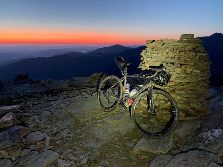 Gravel bike with bikepacking bags beside a stacked-stone cairn under a deep orange twilight sky in the Pyrenees.