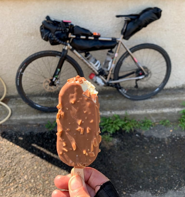 Hand holding a half-eaten nut-coated chocolate ice cream in front of a loaded gravel bike, a hot-day stop during Further.