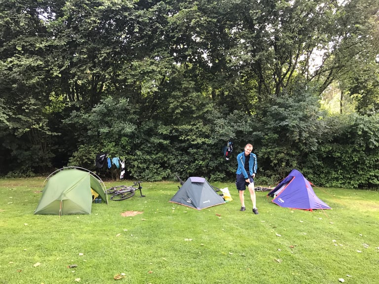 Three small tents on a grassy campsite beneath trees, with a camper standing between them and clothes drying.