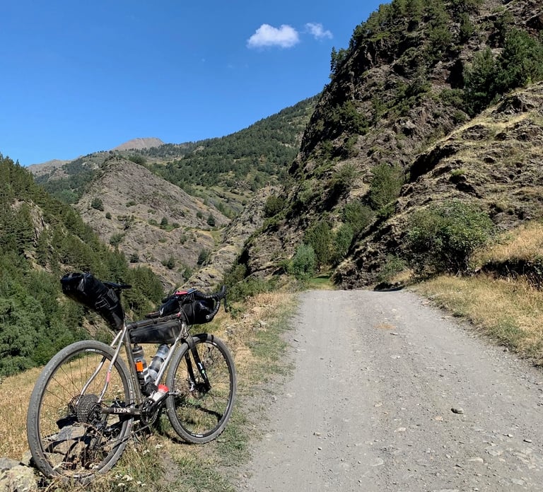 Loaded gravel bike beside a wide gravel road climbing through a rocky, pine-lined Pyrenees valley on the Further route.