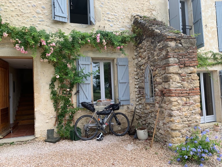 Loaded gravel bike leaning against a stone farmhouse wall with climbing roses, before the Further race in the Pyrenees.