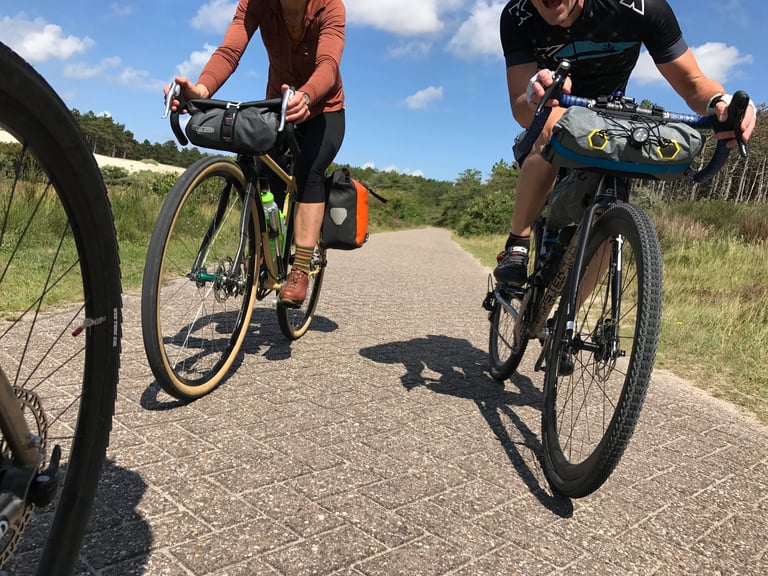 Two riders with loaded bikes on a paved cycle path through dunes and scrub on a sunny day.