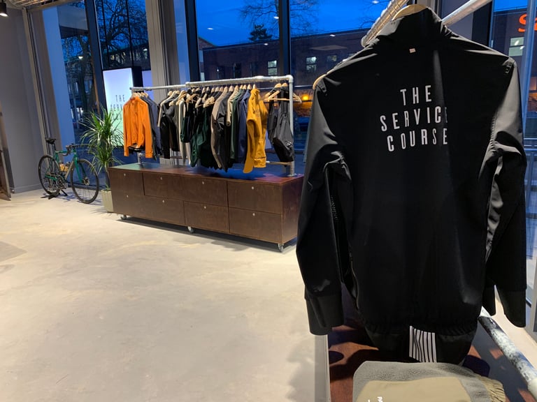 Black jacket printed with The Service Course hangs in front of a clothing rail inside the new Wilmslow store.