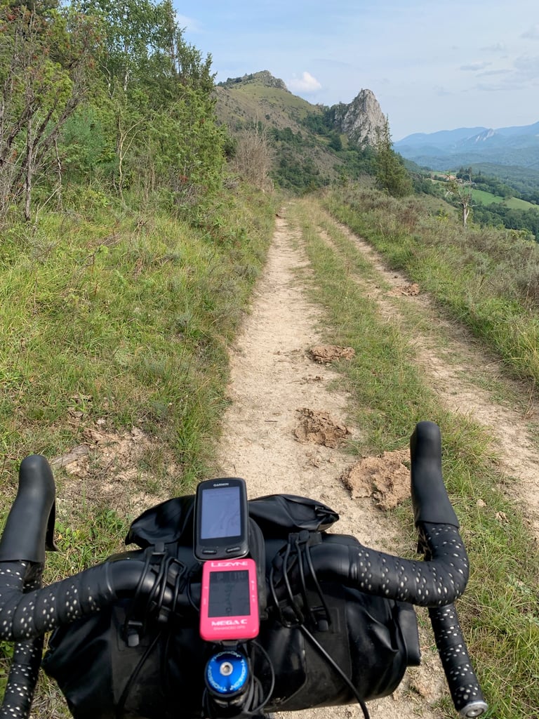 Narrow grassy track climbing towards a rocky crag, seen over Garmin and Lezyne GPS units on a bikepacking bar.
