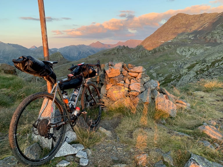 Loaded gravel bike propped on a post beside a stone cairn in evening light on a high Pyrenees pass during Further.