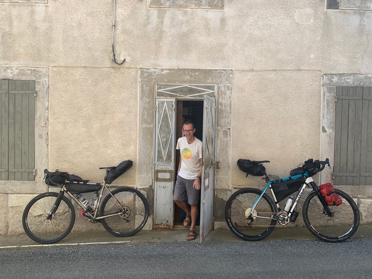 Smiling man in a doorway between two loaded gravel bikes in a French village before the Further race.