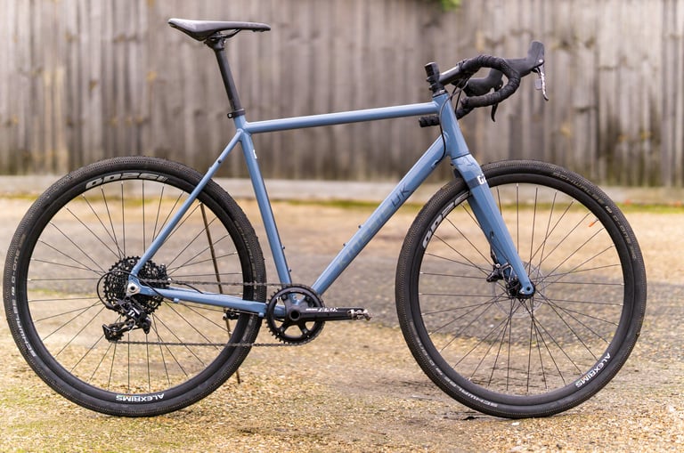Kinesis G2 in slate blue on a gravel yard, with flared drop bars, single-ring SRAM Apex and 38c tyres.