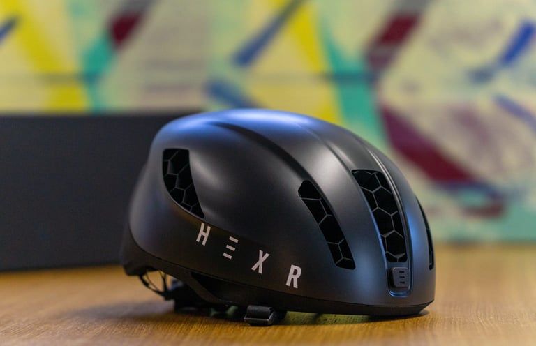Front three-quarter view of the matt black HEXR helmet on a wooden surface, with 3D-printed honeycomb vents.
