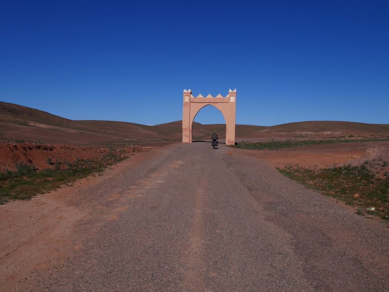 A lone rider with a loaded bike heads towards a pink gateway arch on a gravel road in the Moroccan Atlas.