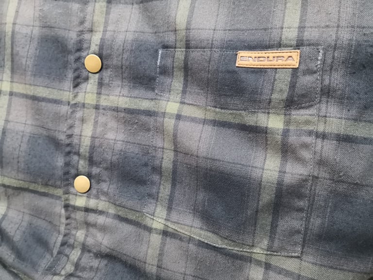 Close-up of the Endura Hummvee Shacket's grey-green plaid side with brass press studs and a leather brand tab.