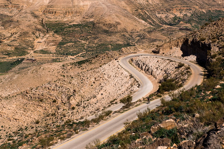 A lone cyclist rides a switchbacking tarmac road through arid, terraced hills on the Jordan Bike Trail.