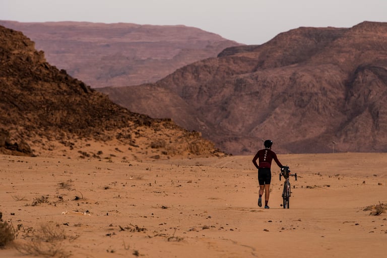 Rider pushes a gravel bike across the sand of the Wadi Rum desert in Jordan, red rock mountains behind.