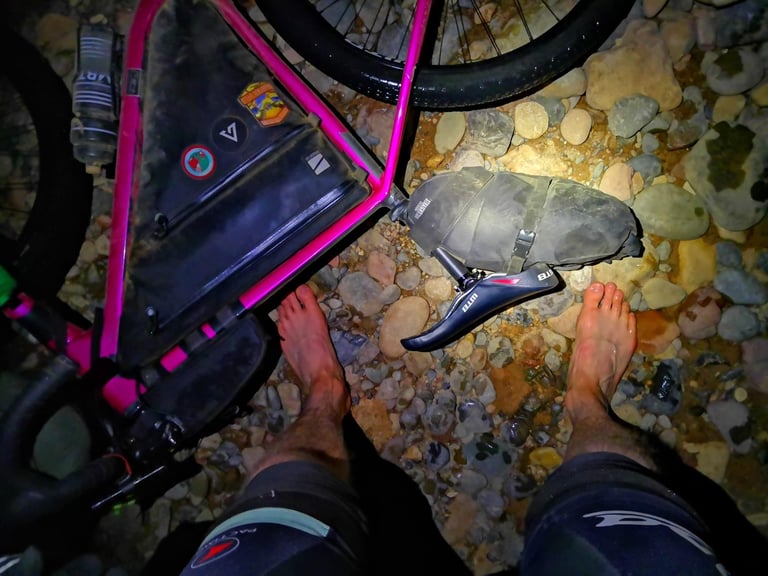 Rider's bare feet on river stones beside the pink Salsa Cutthroat during a night river crossing in Morocco.