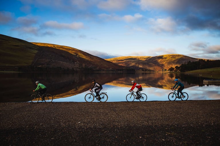 Four cyclists ride along the shore of St Mary's Loch, with golden hills reflected in the still water.